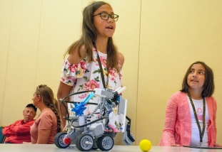 two girls stand above a robot they're getting ready to operate