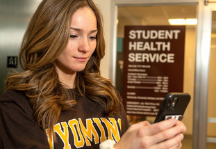 A UW student stands in front of the UW Student Health Services sign as they type on their phone.