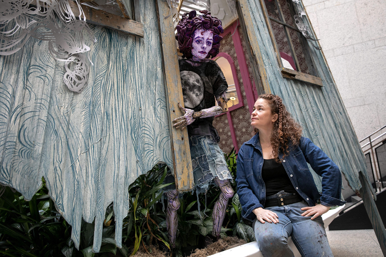 Artist looking up at an art exhibition of a lifesized puppet peering out from behind a door frame.