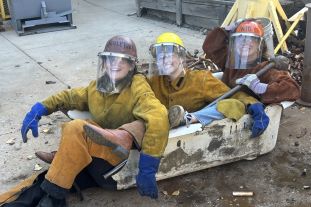 Three students in welding jackets and helmets with facemasks sit in a broken bathtub.