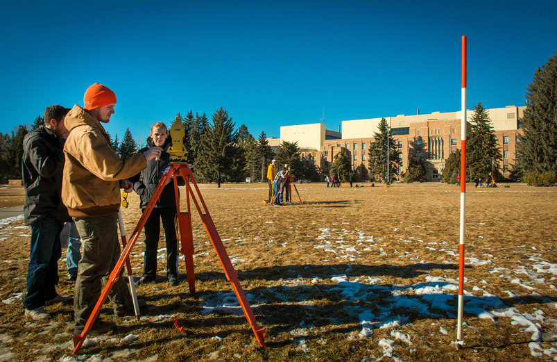 Groups of students practice engineering surveying on Prexy's Pasture. In the foreground, four students stand around a total station while one of them is taking measurements.