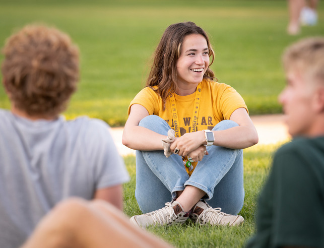 A group of students sit in the grass in Prexy's Pasture while one student looks off to the side and is smiling