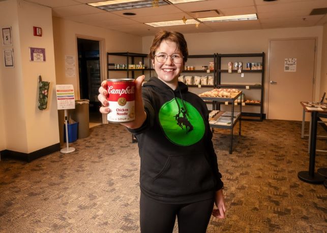 Food Security Coordinator holds up a can of soup in front of the food pantry shelves