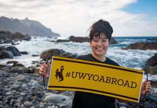 Student holds a UWYO Abroad sign near the ocean