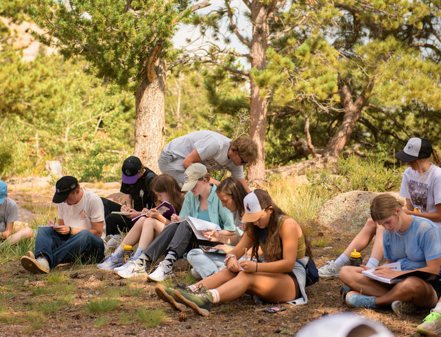 A group of students sit together outside in a forest setting.