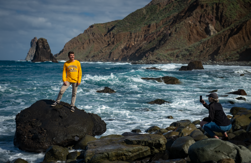 student standing on rock near the ocean while another student takes a photo