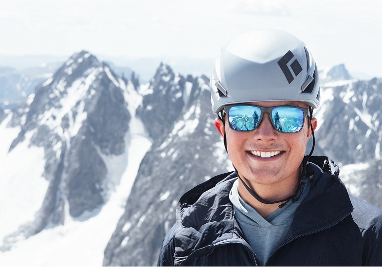 Student wearing a UW yellow shirt sitting on a rock in front of a rocky, mountain landscape with one person sitting in the background.