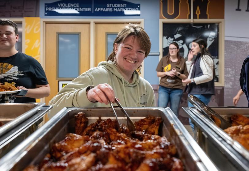 WIHR welcome back party Smiling person serves glazed chicken from a buffet tray while others stand in the background.