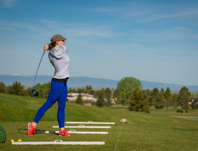 A golfer in blue pants follows through on a golf swing in the backdrop of the snowy range.