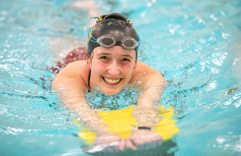 A girl swimming using a foam board and smilling at the camera.