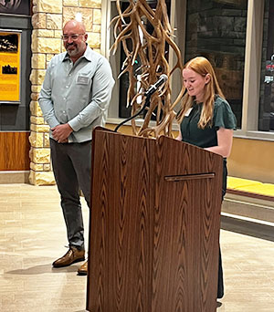 woman speaking from behind a podium while a man stands nearby