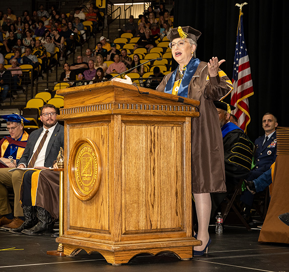 A woman stands behind a podium wearing a cap and gown