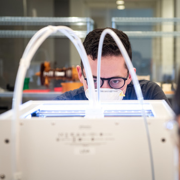 A student peers over a 3D printing machine while wearing a safety mask.