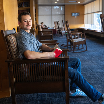 A student smiles for a picture in a rocking chair while holding a book in the library