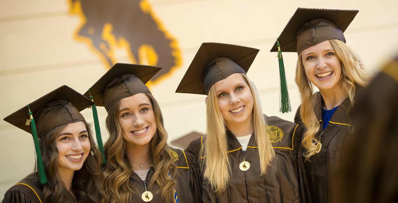 Four UW graduates stand in front of a steamboat insignia and smile broadly.