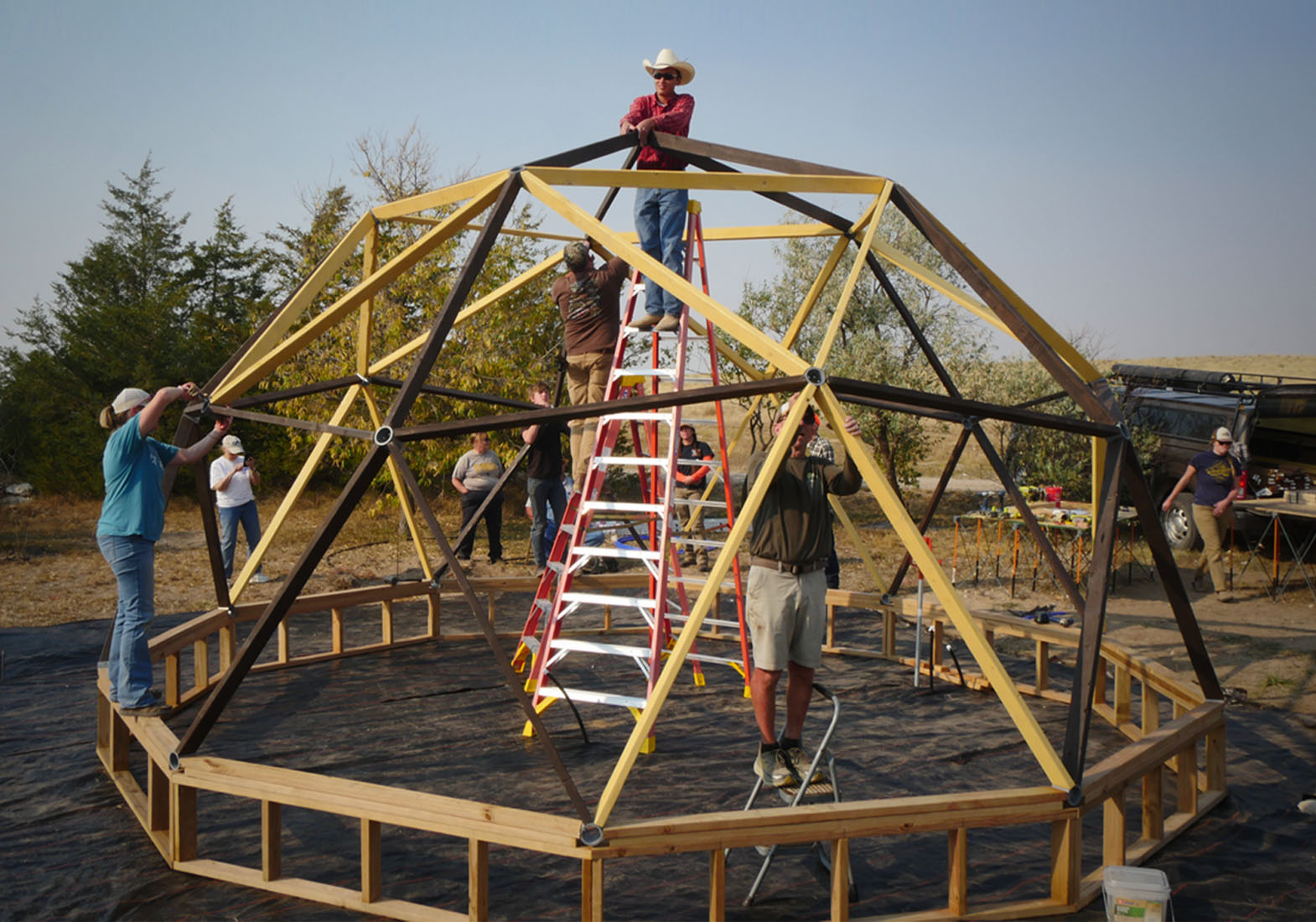 Educators use ladders to assemble the wooden frame of a geodesic dome outdoors