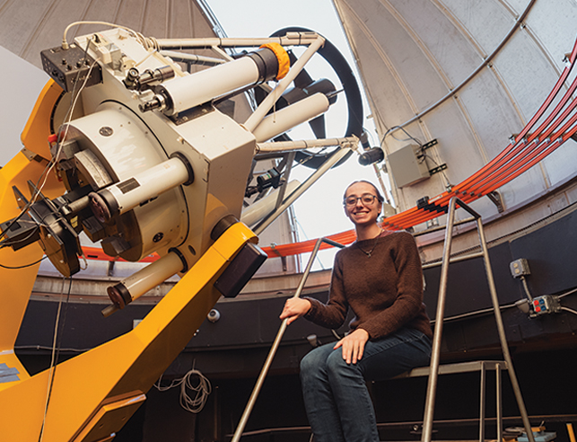 female college student sitting by large telescope