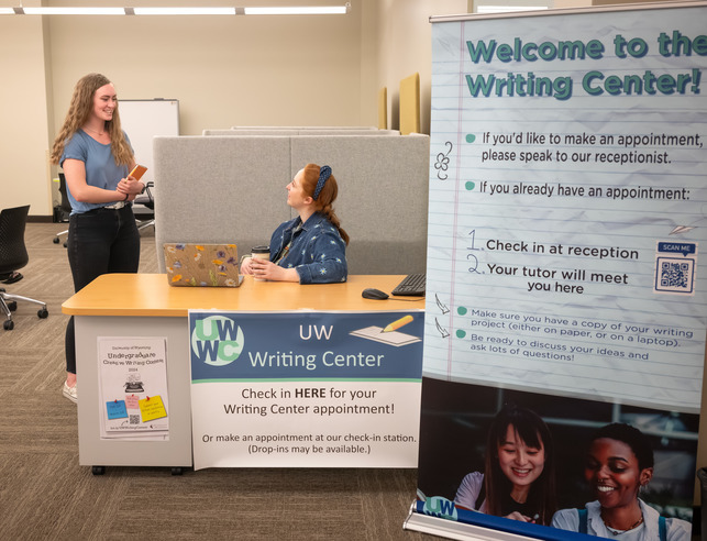 The reception area of the writing center with the welcome sign present and two students speaking at the reception desk